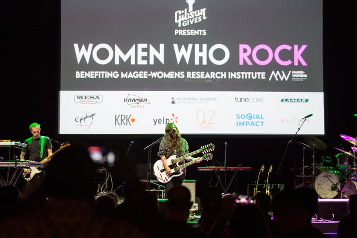 Musicians perform on stage at the 'Women Who Rock' event, with a large screen in the background displaying sponsor logos and the event's name.