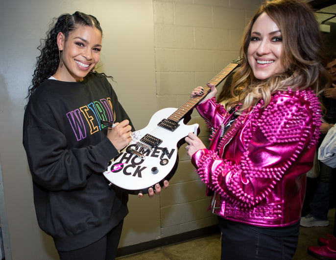 Jordin Sparks signs a white 'Women Who Rock' guitar backstage while Melinda Colaizzi, wearing a pink studded jacket, smiles beside her.