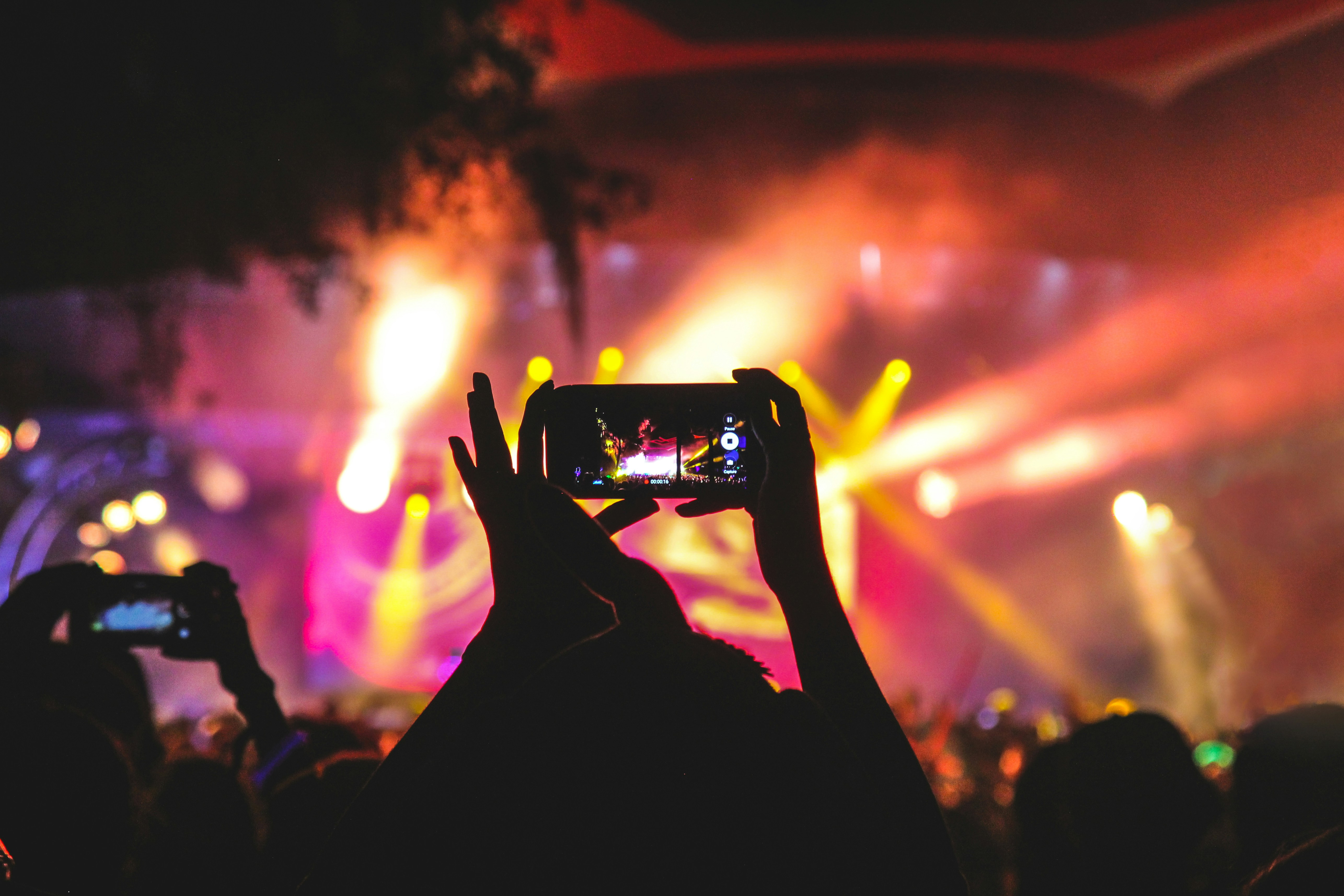 Silhouette of a person holding up a smartphone to capture a live concert, with vibrant stage lights and colorful smoke in the background. The bright yellow, pink, and orange lights create a dynamic and energetic atmosphere, emphasizing the excitement of the live event.