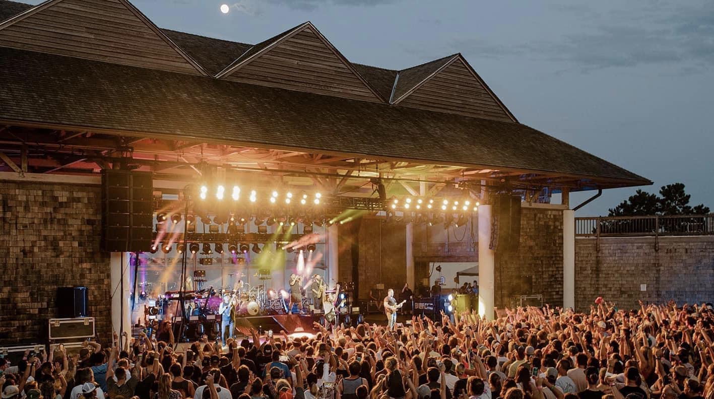 A vibrant evening concert with bright stage lights and a large crowd gathered at an outdoor venue.