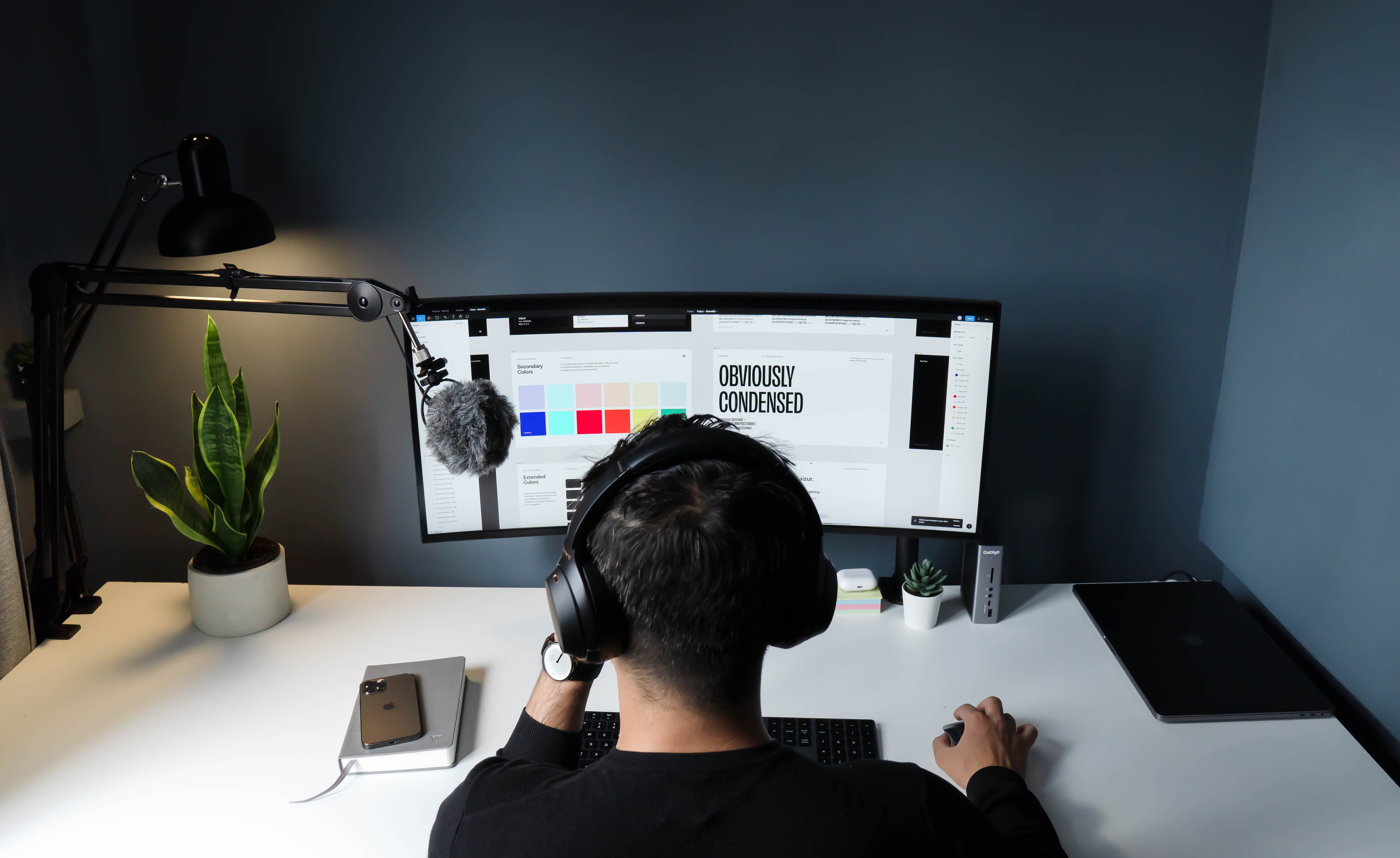 Person working at a desk with a dual-monitor setup, viewing a design project on the screen that includes color swatches and typography labeled 'OBVIOUSLY CONDENSED.' The workspace features a desk lamp, microphone on a boom arm, headphones, potted plants, a smartphone, and a laptop, creating a modern and organized home office environment.