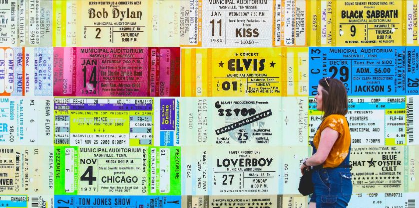 Collage of vintage concert tickets featuring various famous artists like Bob Dylan, Elvis, Kiss, Black Sabbath, and Jackson 5, among others. The tickets are arranged in a colorful, overlapping pattern, creating a nostalgic tribute to past concerts. A person stands on the right side, looking at the tickets, wearing a yellow shirt and denim overalls, adding a contemporary element to the retro-themed display.