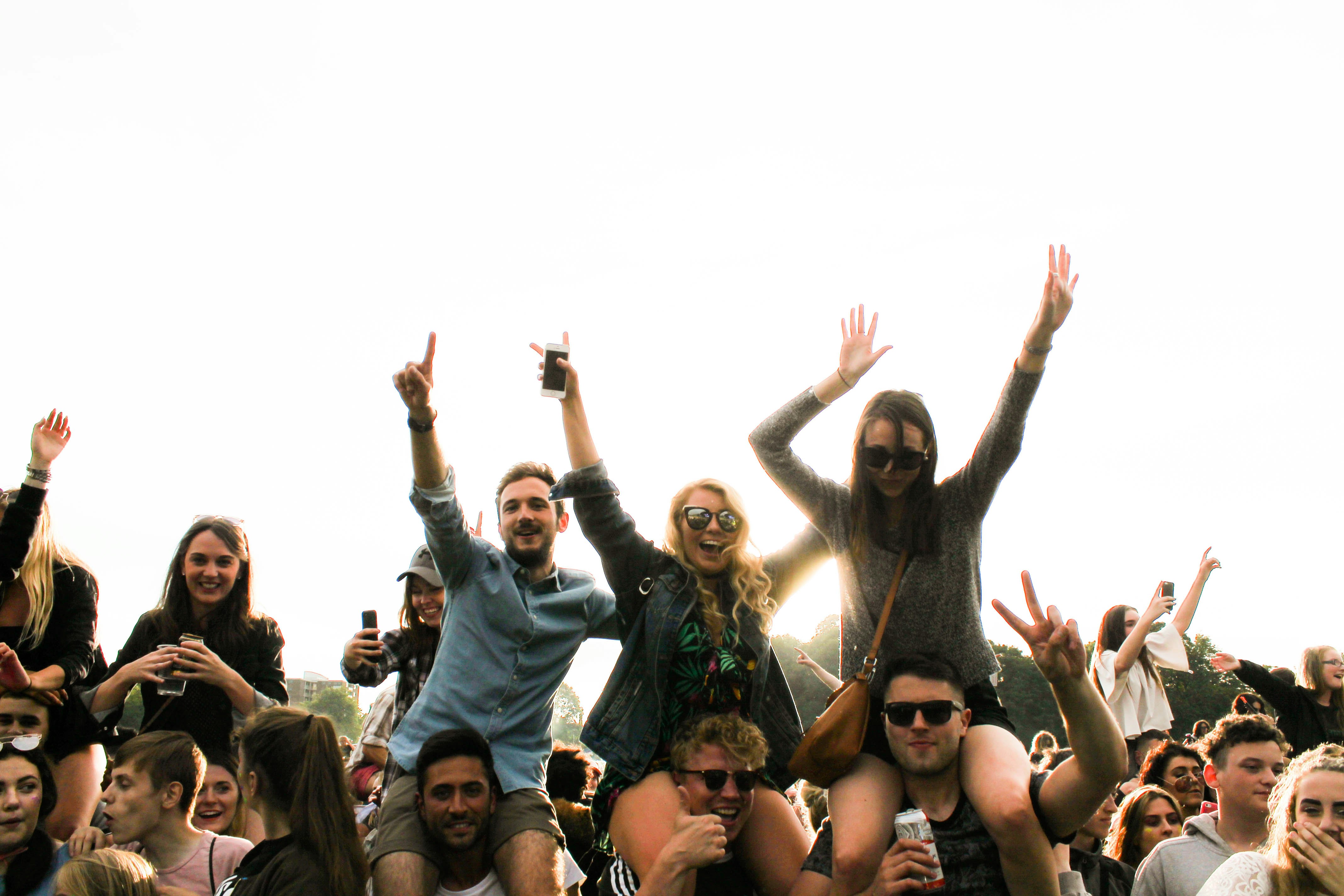 Group of young people at an outdoor event, enjoying a lively atmosphere. Some individuals are sitting on others' shoulders, raising their arms, and smiling. The crowd is diverse, with people holding drinks and capturing moments on their phones, showcasing a fun and energetic festival or concert vibe.