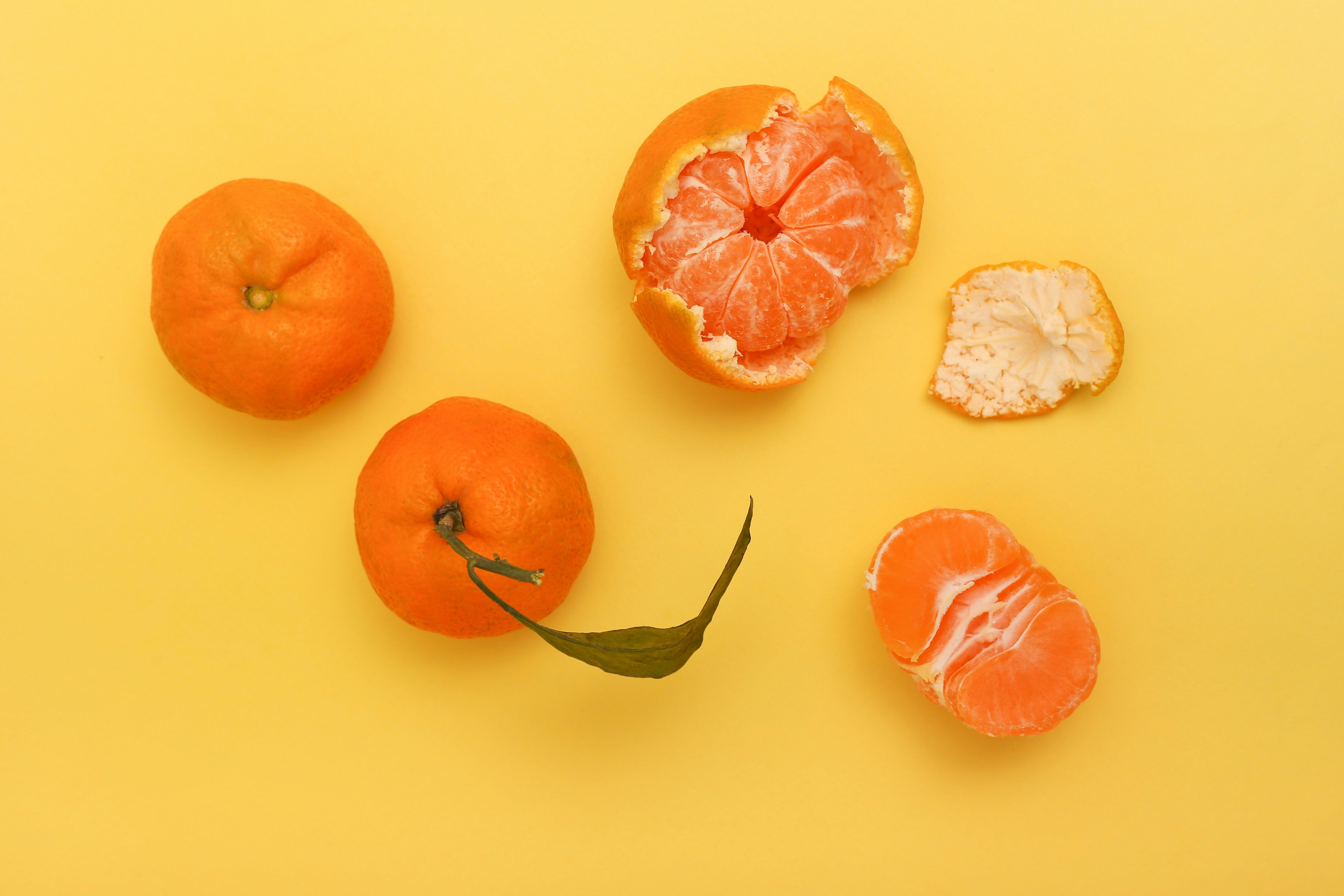 Several mandarins on a yellow background, including whole, partially peeled, and fully peeled segments. One mandarin has a leaf attached, and the bright citrus fruits are arranged artistically to showcase their vibrant orange color against the contrasting yellow surface.