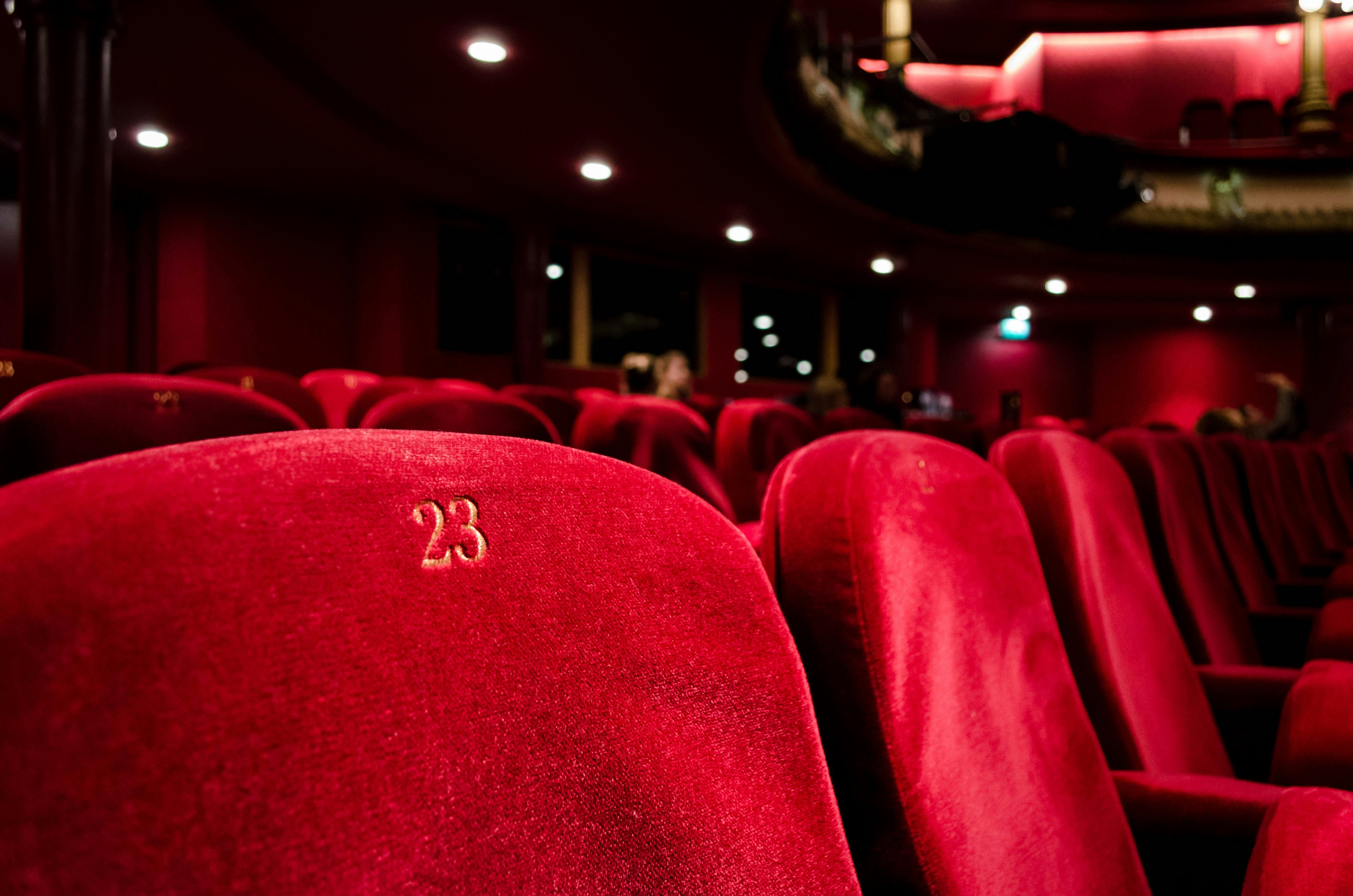 Close-up view of plush red theater seats with the number '23' embroidered on one of the seats. The theater setting is dimly lit, creating a warm and intimate atmosphere, with soft lighting and the hint of a balcony in the background. The focus is on the rich texture and vibrant color of the seats, highlighting the classic elegance of the venue.