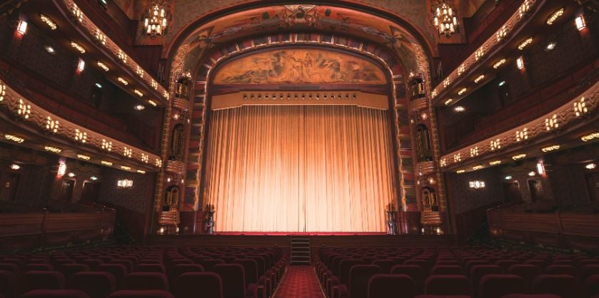 Interior view of a grand theater with an ornate, classic design. The image features a large stage with a closed, illuminated curtain, surrounded by intricate architectural details, including multiple balconies with rows of seats and decorative lighting. The warm lighting creates an elegant and nostalgic ambiance, highlighting the theater's rich and traditional decor.