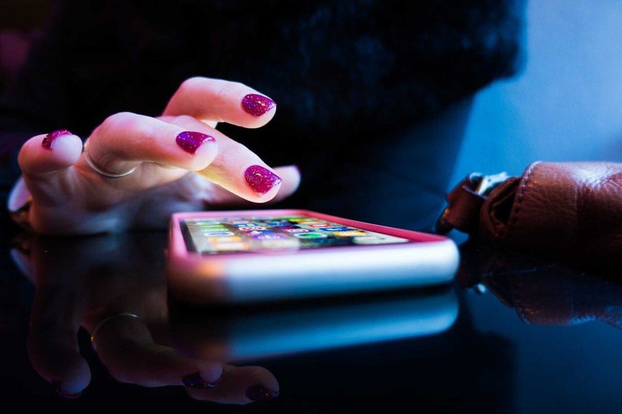 Close-up of a hand with glittery purple nail polish tapping on a smartphone screen. The phone lies on a reflective surface with a leather purse nearby, illuminated by soft blue lighting, creating a tech-focused, stylish atmosphere.