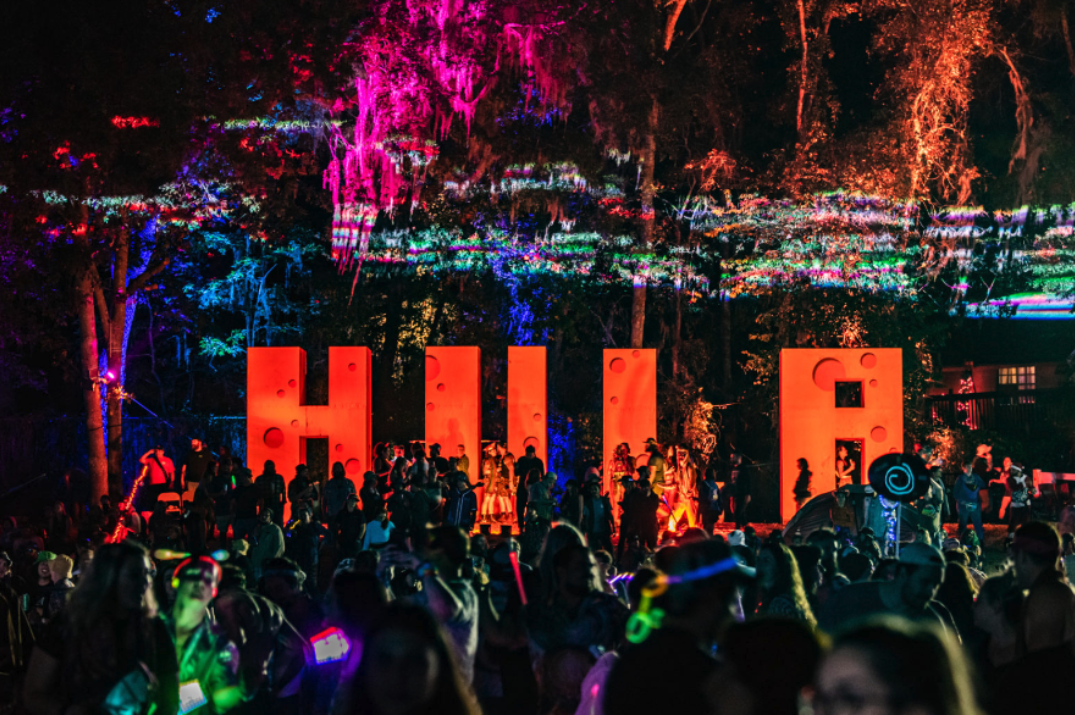 Colorful nighttime crowd at the Hulaween festival, with large illuminated 'HULA' letters and vibrant lighting effects in the surrounding trees.