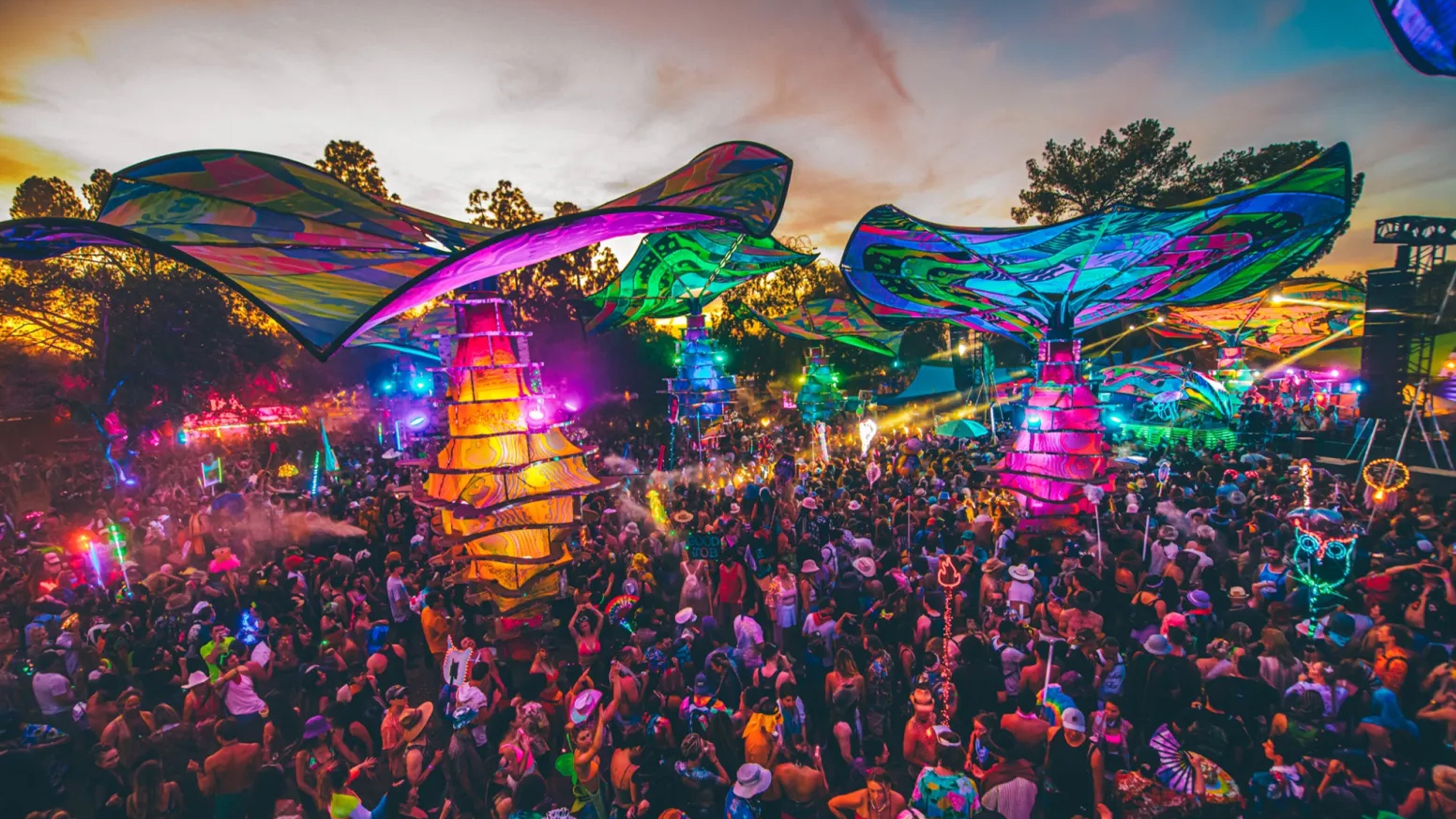 A vibrant outdoor festival scene at sunset, filled with a large crowd of attendees under colorful, illuminated canopy structures. The atmosphere is lively, with vivid lights, artistic decorations, and people dressed in festival attire, creating a dynamic and energetic environment.