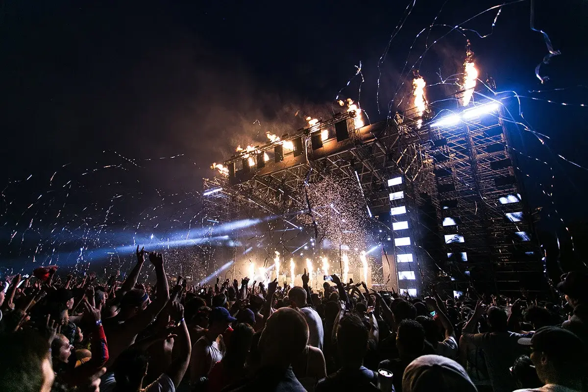 Nighttime crowd at Vibra Urbana festival cheering in front of a stage with pyrotechnics, confetti, and vibrant lighting effects.